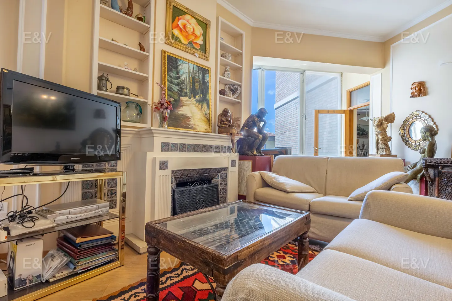 Living room with beige walls, white built-in shelves, and a fireplace. A TV sits on a gold stand, and a glass-topped coffee table is on a patterned rug.