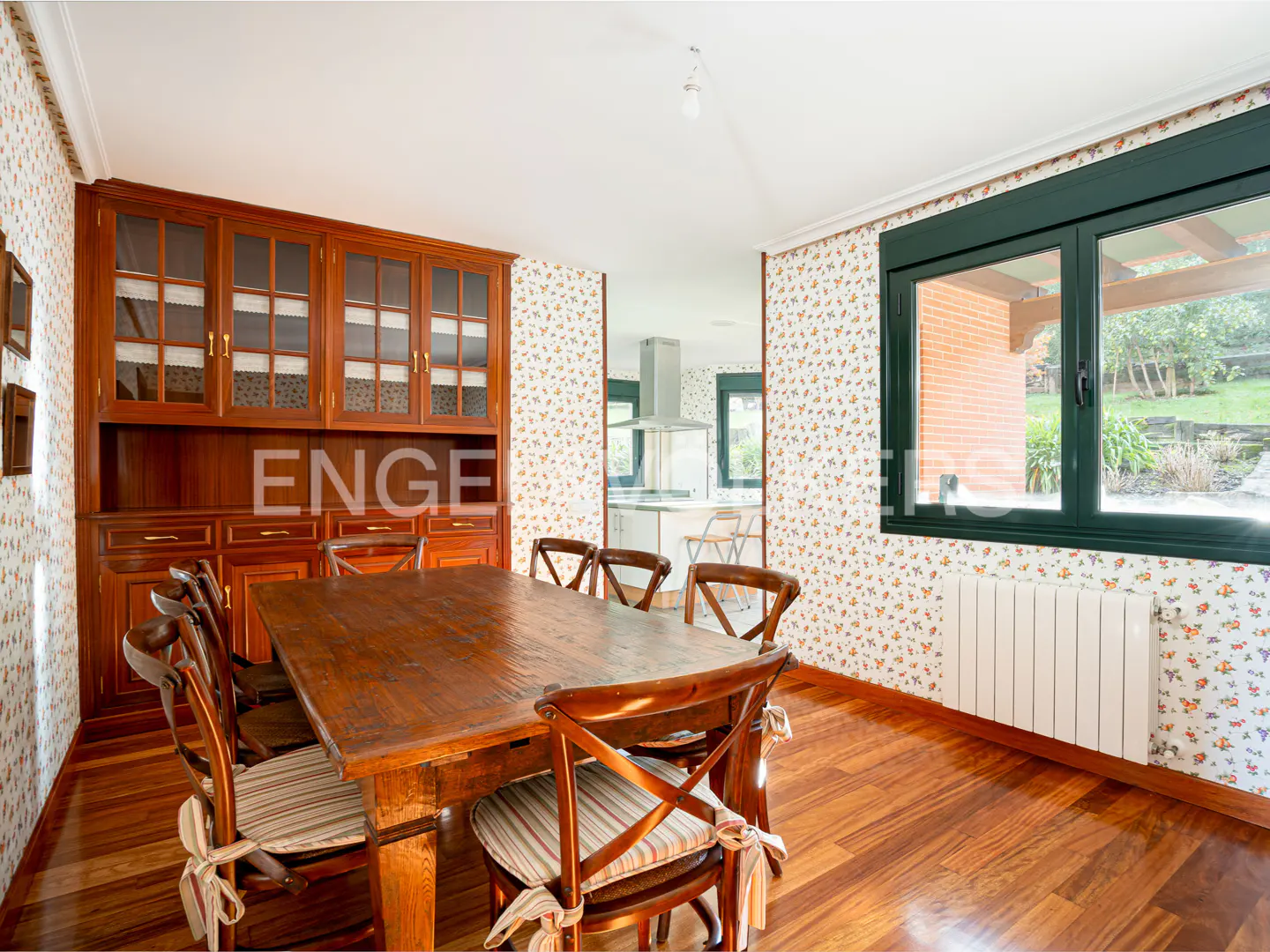 A dining room with a wooden table, chairs, and a built-in cabinet. Floral wallpaper and a window with a view of the garden.