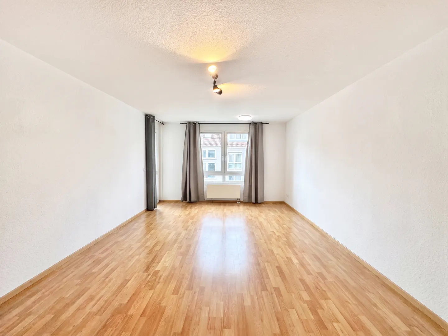 Bright, empty room with hardwood floors, white walls, and a window with gray curtains.