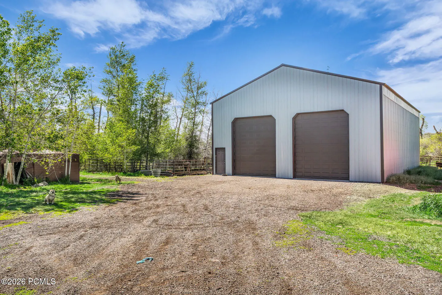 Large metal workshop with two brown garage doors on a gravel lot, surrounded by trees and a blue sky.