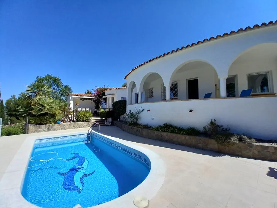 A bright, sunny view of a white villa with arched porch overlooking a blue swimming pool with mosaic design.