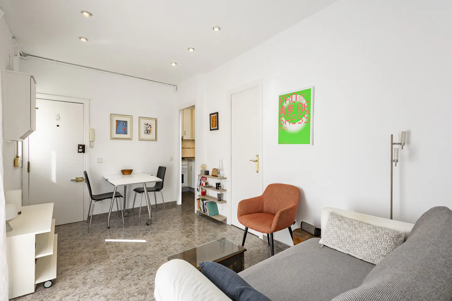 Bright, modern apartment interior with white walls, gray tile floor, and recessed lighting. Features a dining table, orange chair, and gray sofa.