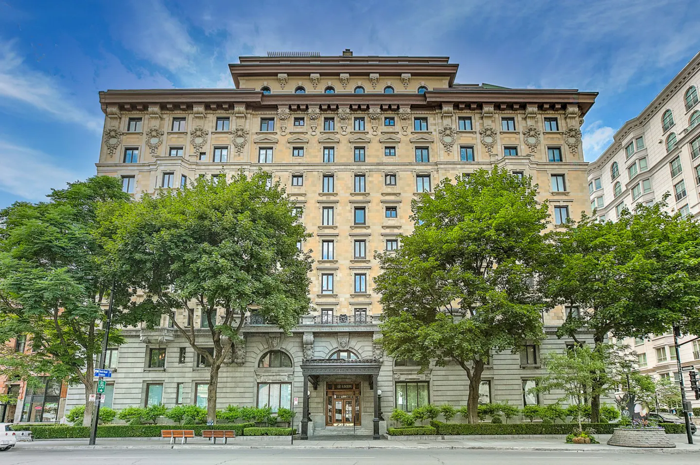 Exterior view of a large, beige apartment building with many windows and ornate detailing, framed by green trees.