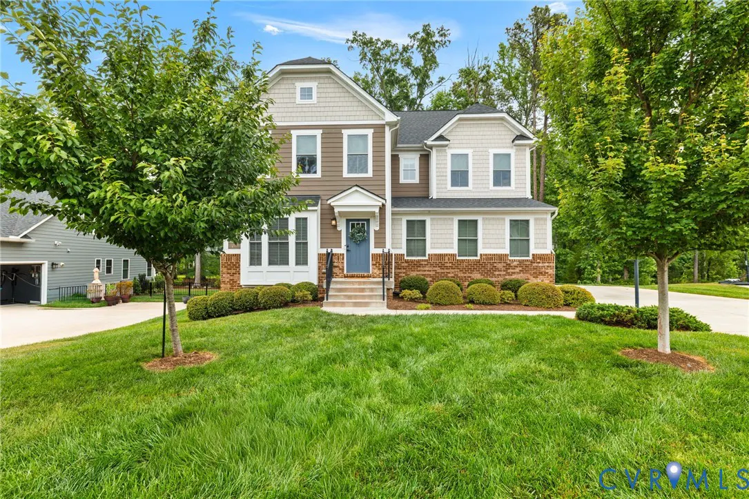 Two-story house with brown and gray siding, brick base, blue front door, green lawn, and trees.
