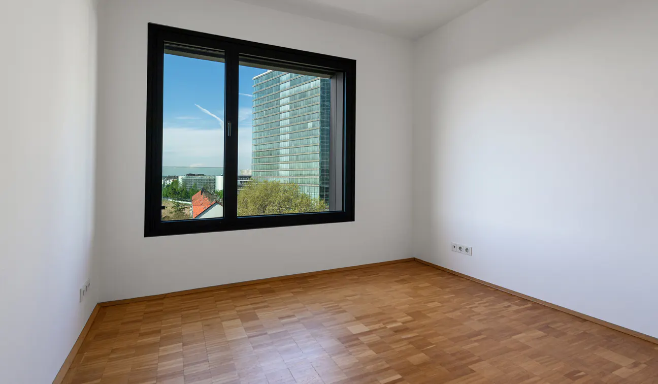 Empty room with white walls, wood floor, and black-framed window showing a city view with a tall building.