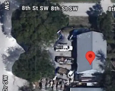 Aerial view of a gray building with a red location pin, surrounded by trees and parked vehicles.