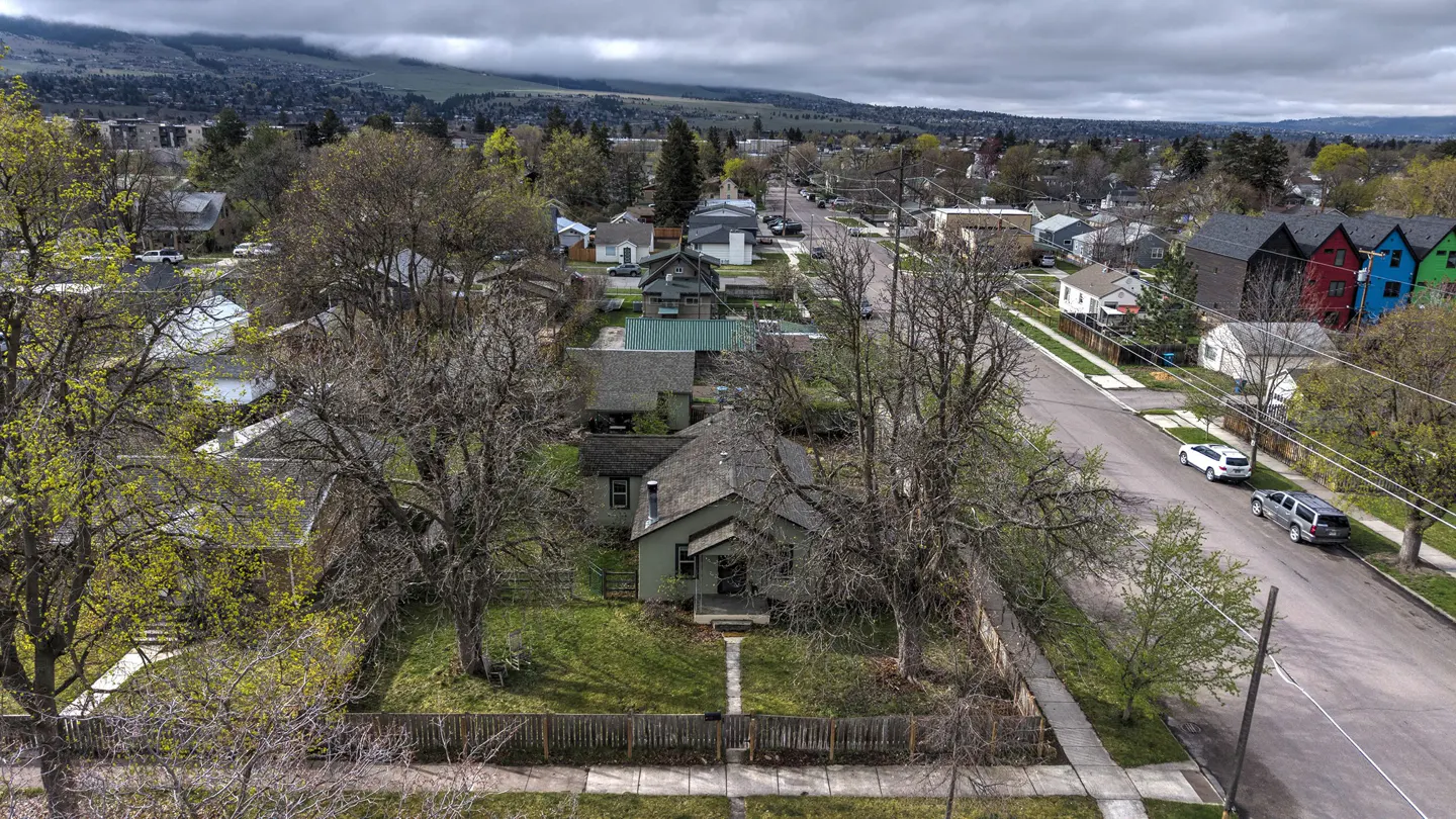 Aerial view of a quiet residential street with houses, trees, and cars. A mountain range is visible in the background.