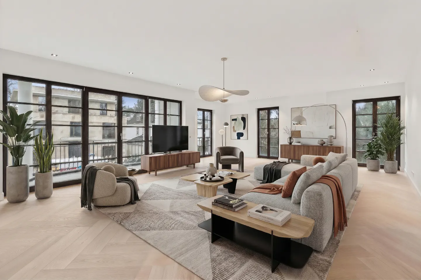 Bright living room with large windows, herringbone floors, and a gray sectional sofa. Modern furniture and potted plants add to the stylish decor.