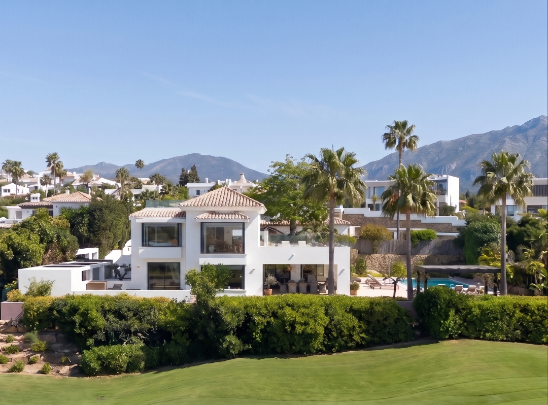 Modern white villa with a tiled roof, palm trees, a swimming pool, and lush landscaping against a mountain backdrop.