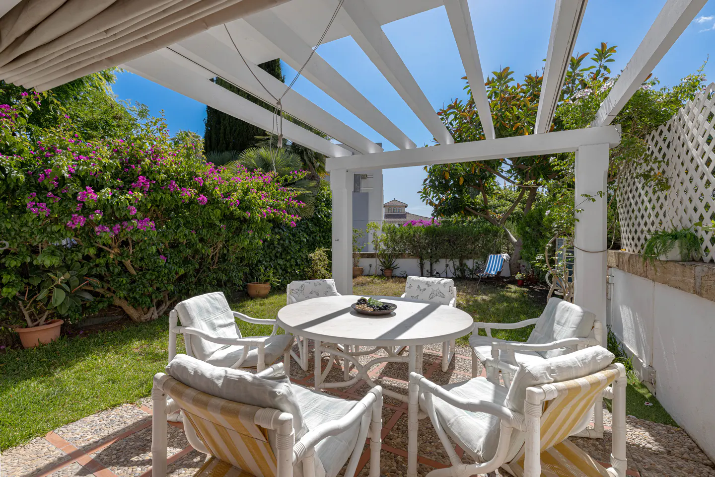 White patio furniture set under a pergola in a sunny garden with vibrant pink bougainvillea and green lawn.