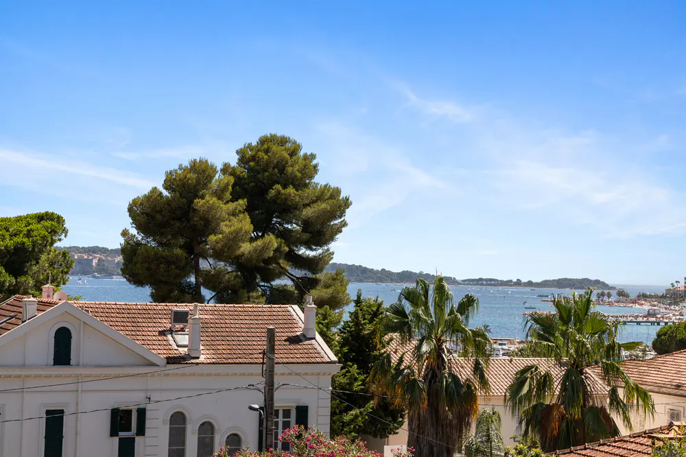 View of a white villa with a terracotta roof, palm trees, and a blue sea under a clear sky in a coastal town.