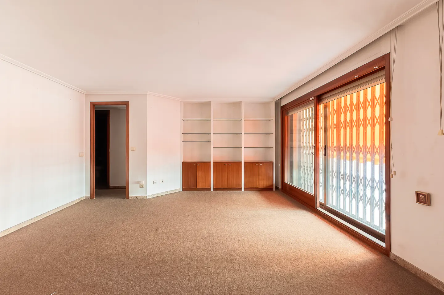 Empty living room with brown carpet, white walls, built-in shelves, and a sliding glass door with orange security bars.