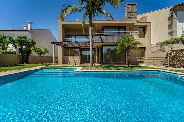 A modern tan house with a blue swimming pool and palm trees on a sunny day.