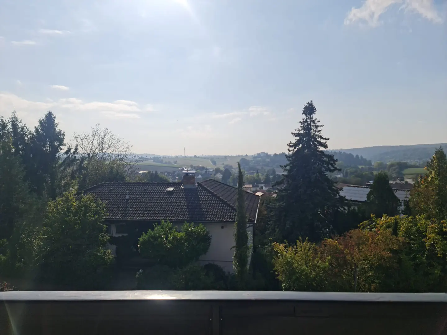 A sunny view from a balcony overlooking a house with a brown tiled roof, green trees, and a distant town.