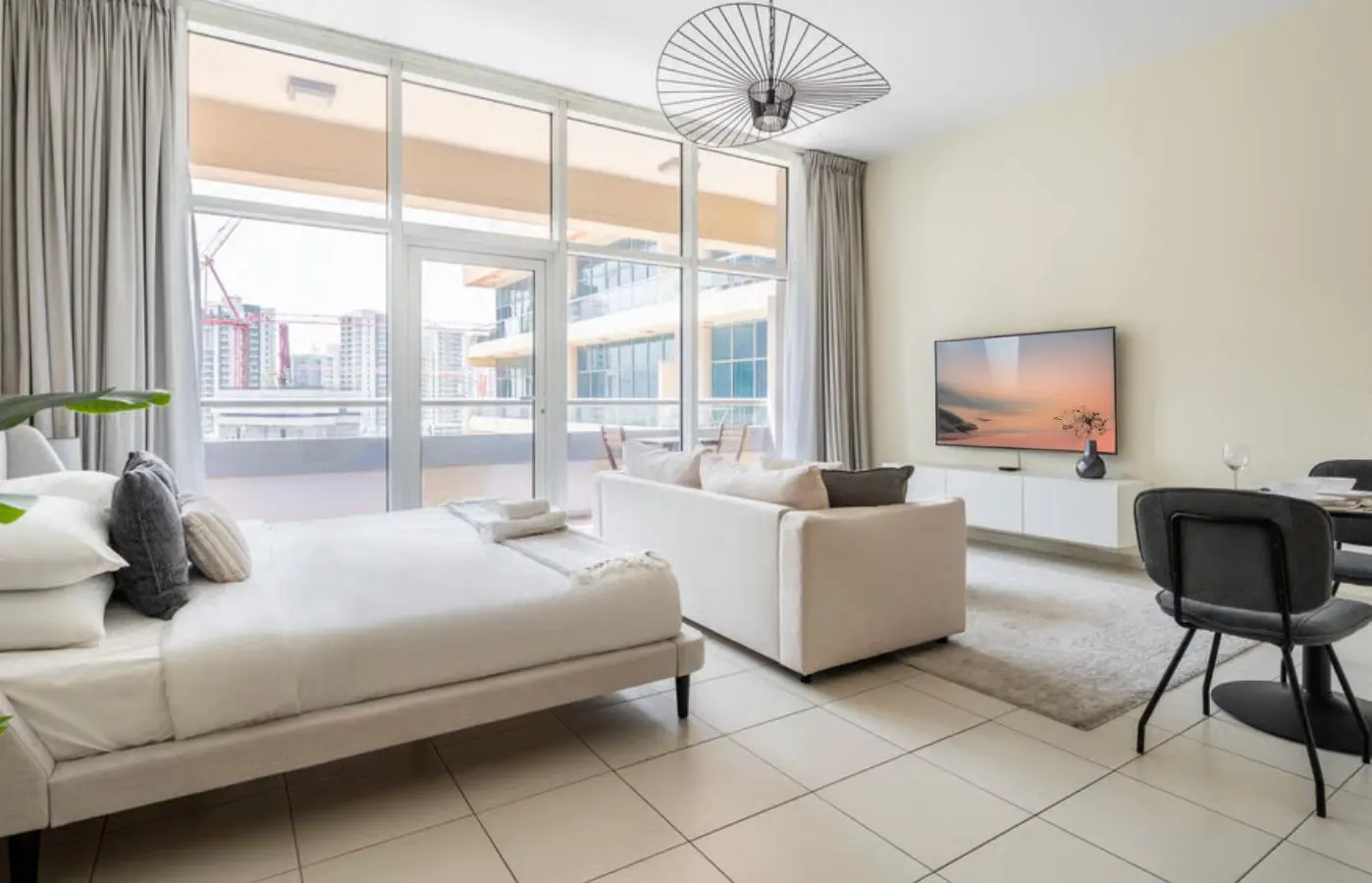 Bright studio apartment with a bed, sofa, TV, and dining table. Large windows offer a city view. Neutral tones create a modern, open space.