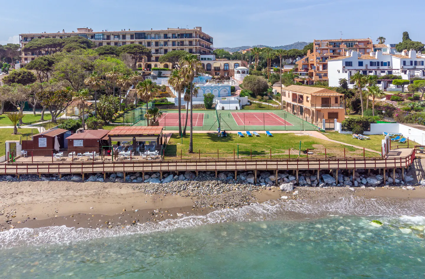 Coastal view of a resort with tennis courts, beachside cabanas, and turquoise water under a blue sky.