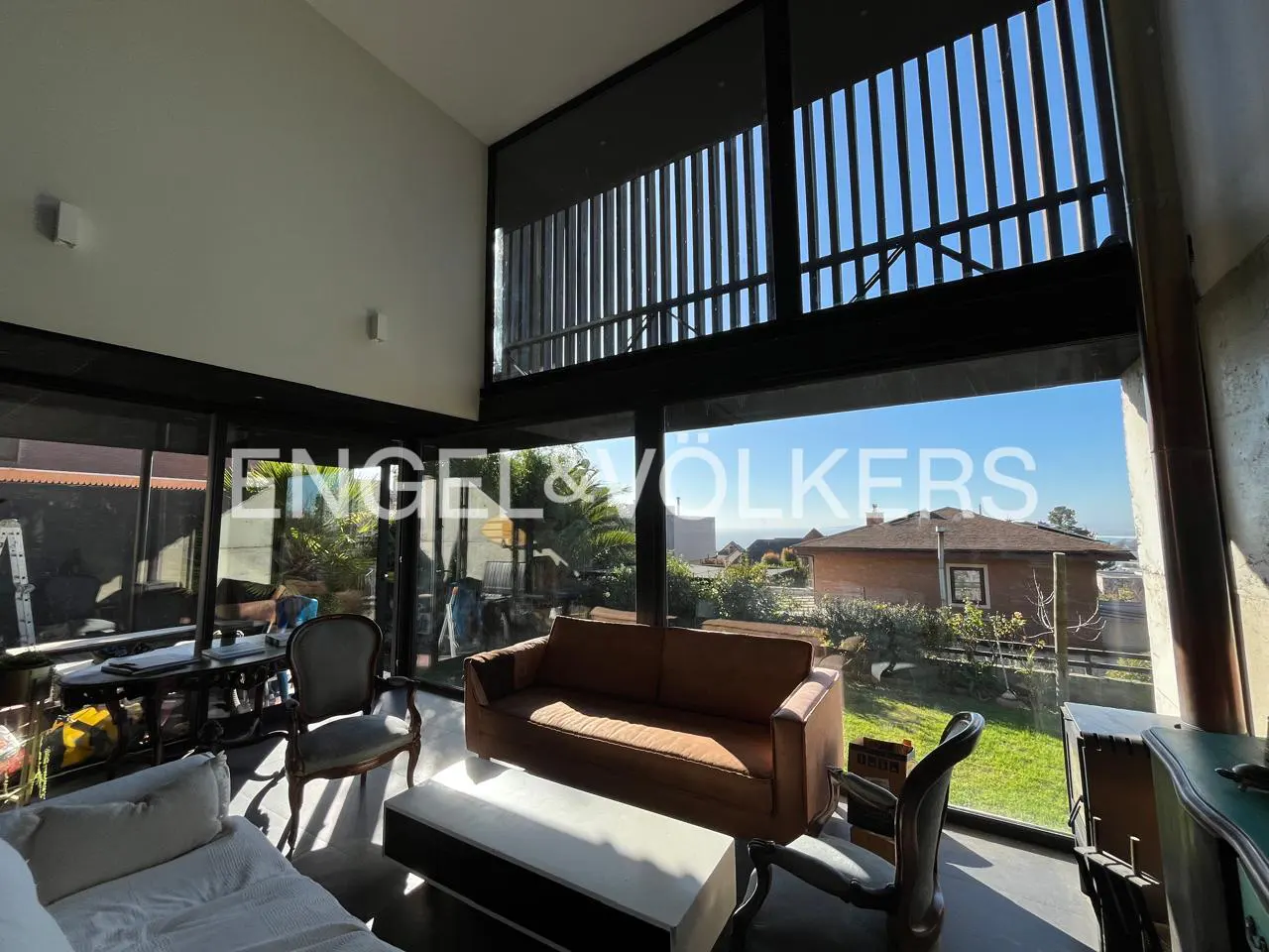 Bright living room with a brown sofa, white coffee table, and large windows overlooking a garden. Engel & Völkers logo visible.