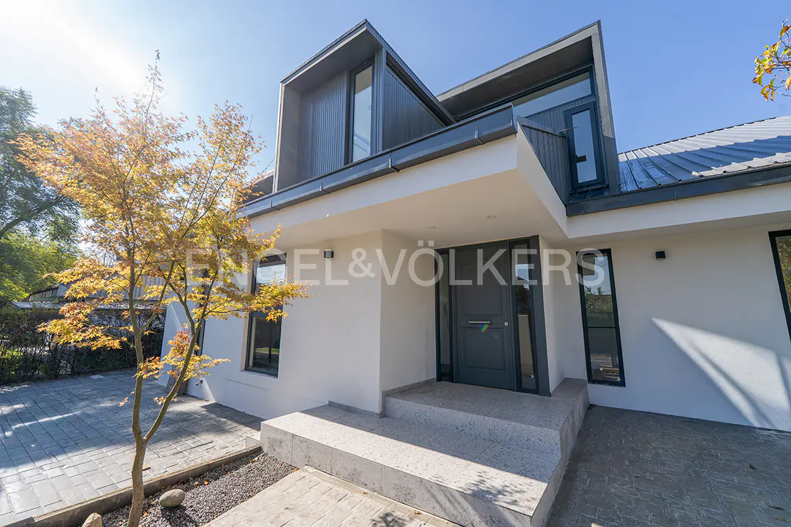 Modern two-story house with a gray door, white walls, and gray trim. A small tree with yellow leaves is on the left. "Engel & Völkers" logo is visible.
