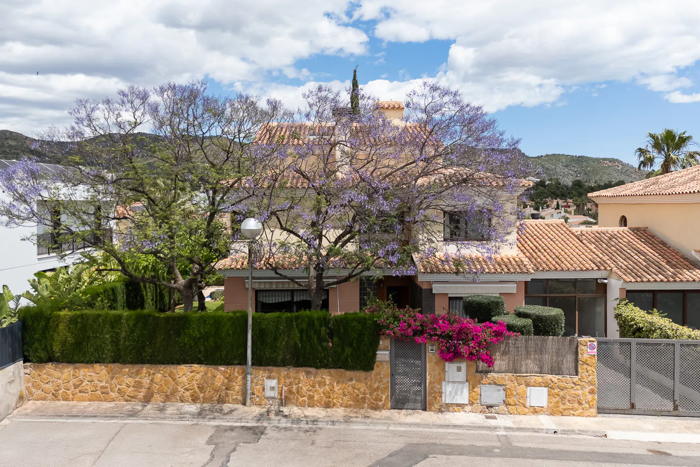 Two-story house with a red tile roof and a large tree with purple flowers in front. A stone wall and green hedge surround the property.