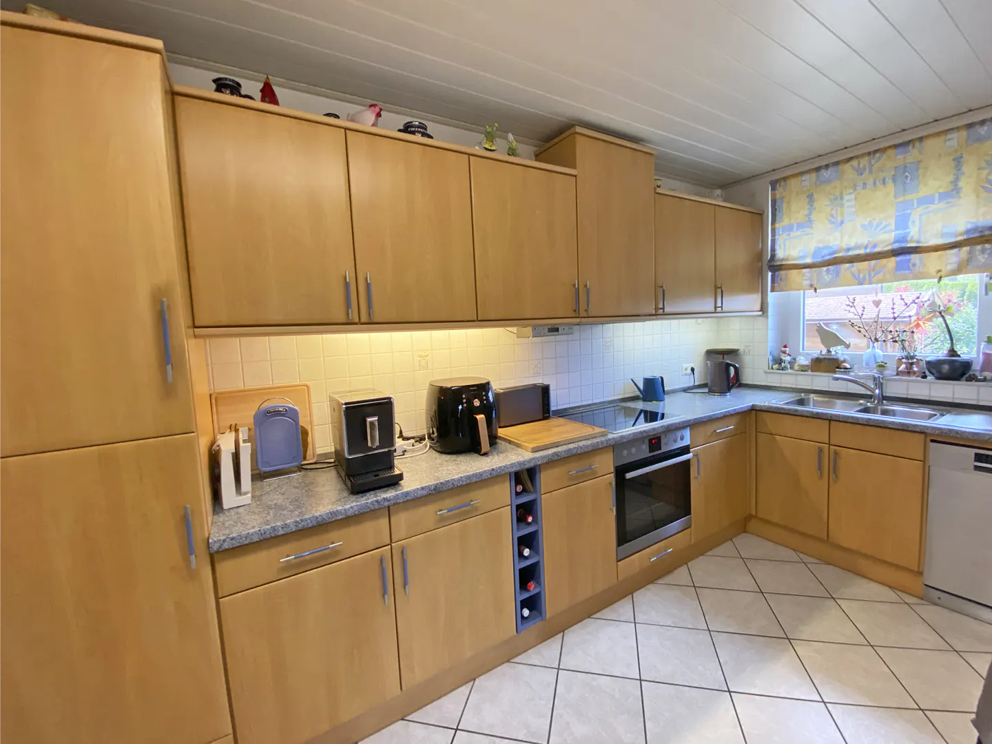 A bright kitchen with light wood cabinets, gray countertops, and white tile floors. A window with a blue and yellow patterned shade is above the sink.