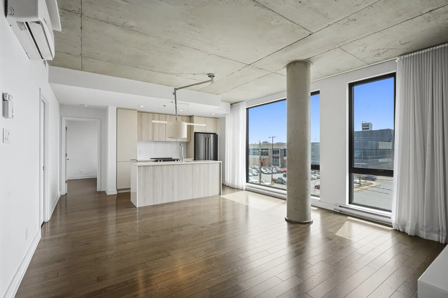 Bright, modern apartment interior with hardwood floors, concrete ceiling, and large windows. Kitchen island and stainless steel appliances visible.