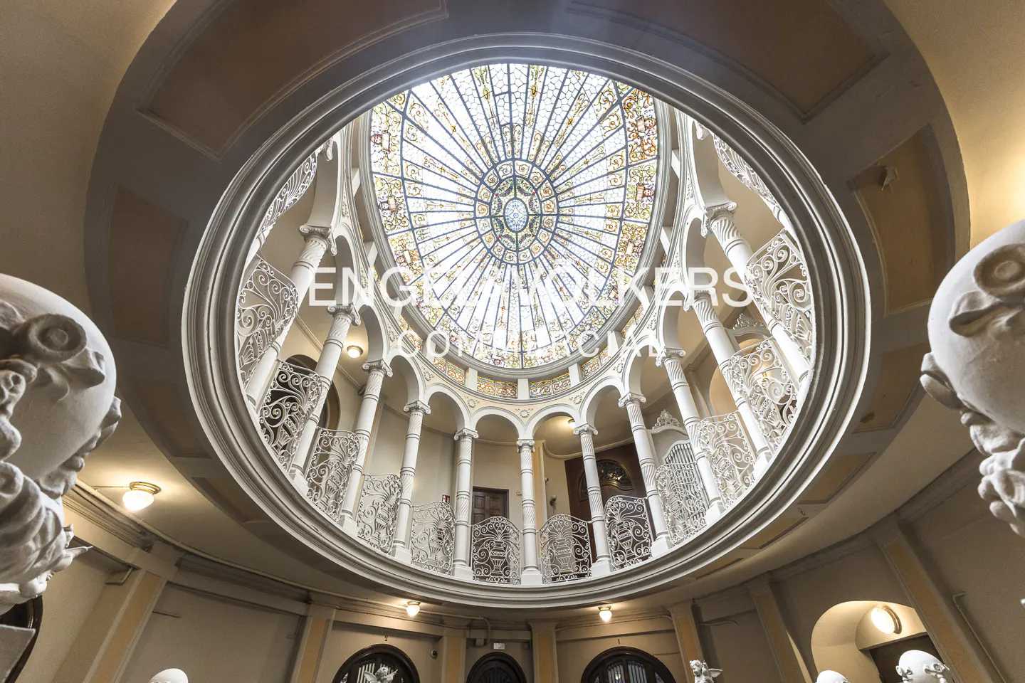 Looking up at a building's interior, featuring a stained-glass dome, white columns, and ornate railings.