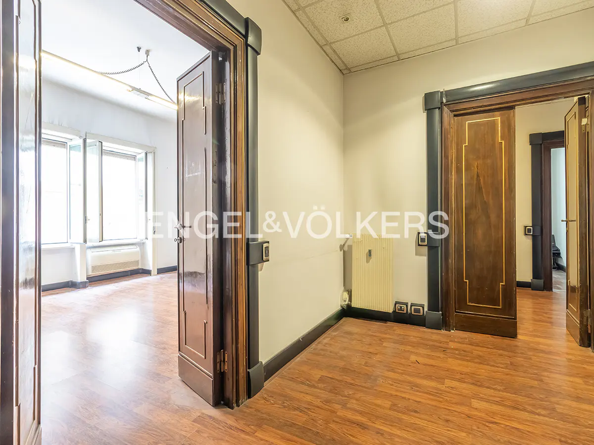 A hallway with wood-look flooring, white walls, and two open dark wood doors leading into bright, empty rooms.