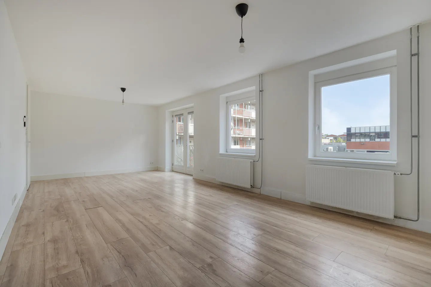 Bright, empty room with wood floors, white walls, and two windows. Radiators are under the windows. Two pendant lights hang from the ceiling.