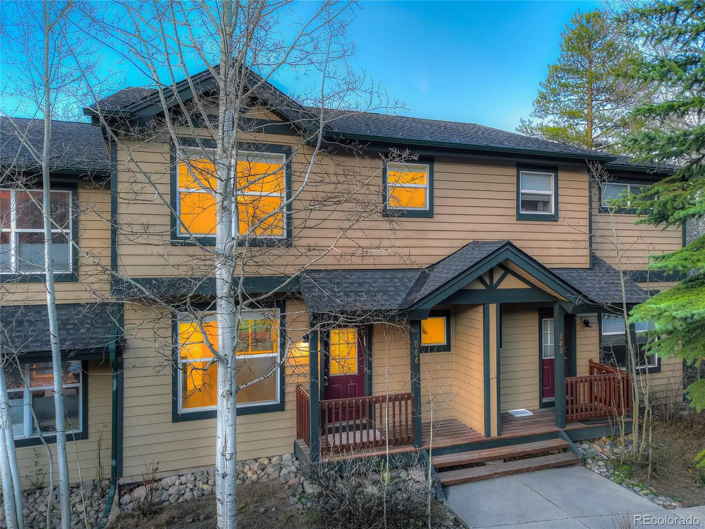 Two-story tan townhouse with dark trim, a dark roof, and a small porch with a red door. Windows glow with warm light.