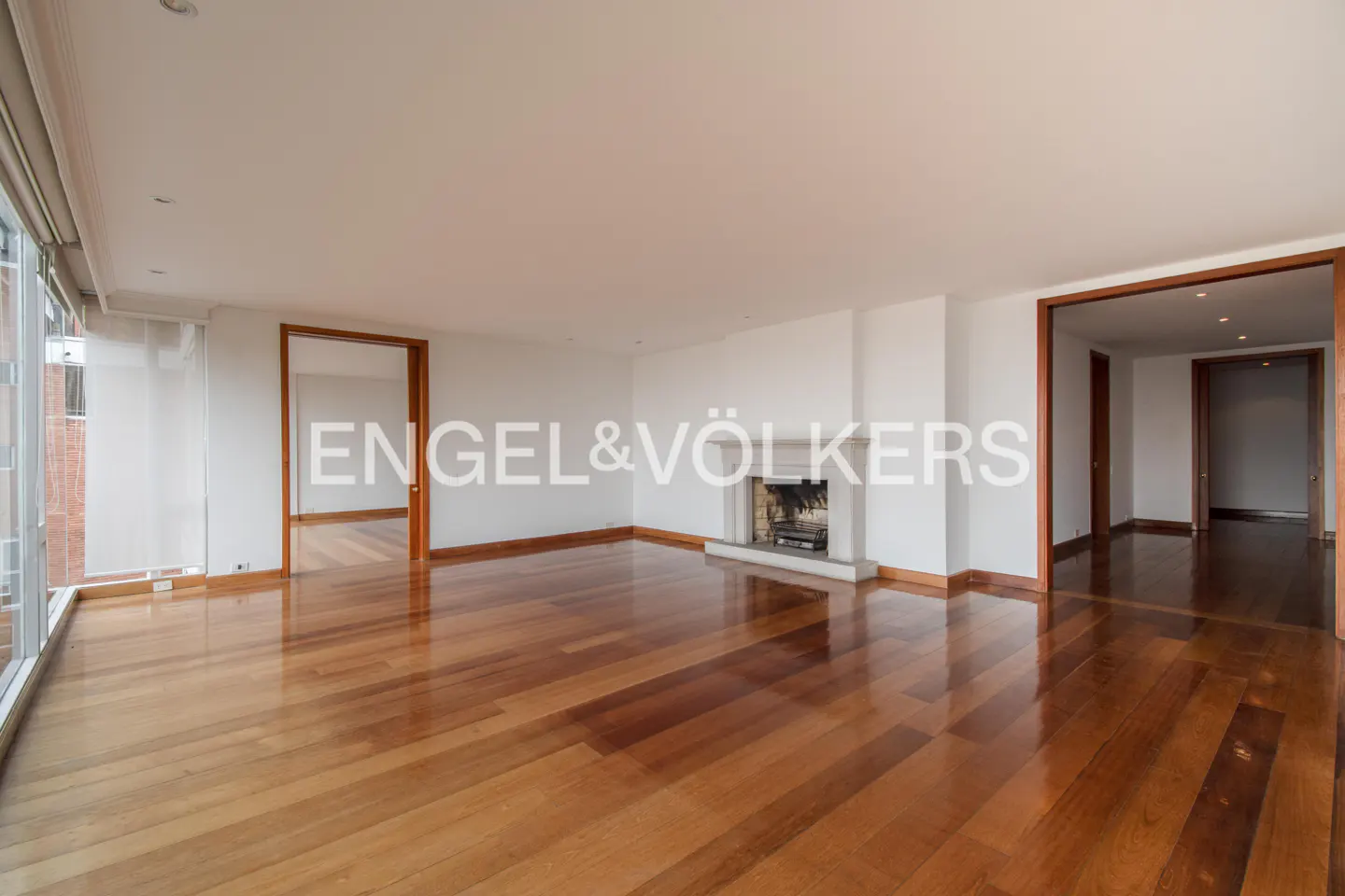 Bright, empty living room with hardwood floors, white walls, and a fireplace. Doorways lead to other rooms. Engel & Völkers logo visible.