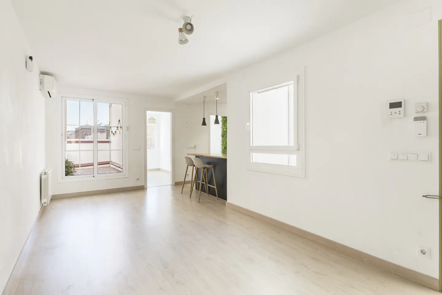 Bright, empty room with light wood floors, white walls, and a window to a balcony. A kitchen bar with two stools is visible.