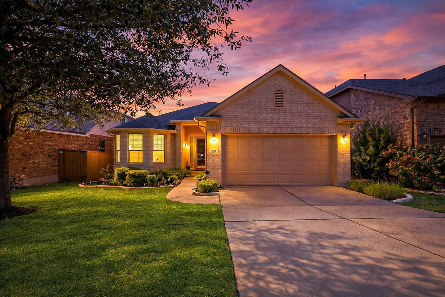 A tan brick house with a two-car garage, green lawn, and a pink and purple sunset sky.