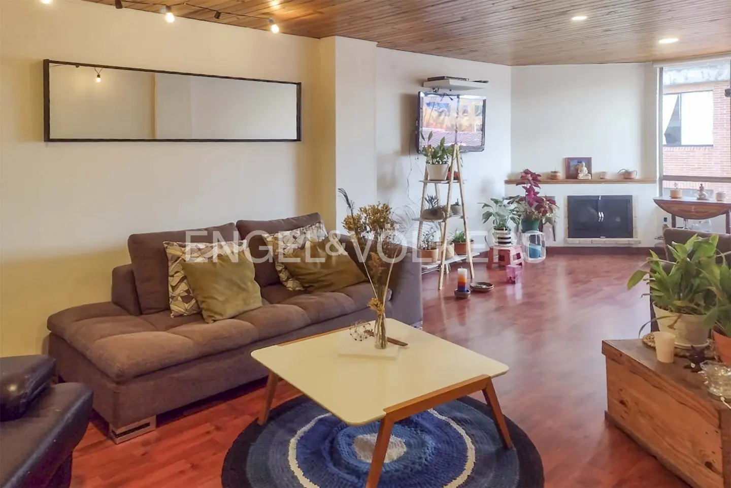 Living room with brown sofa, square coffee table on a blue rug, and a fireplace with plants. Wooden ceiling and hardwood floors.