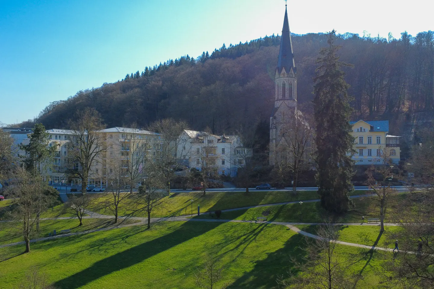 View of a European town with a church steeple, apartments, and a green park under a blue sky.