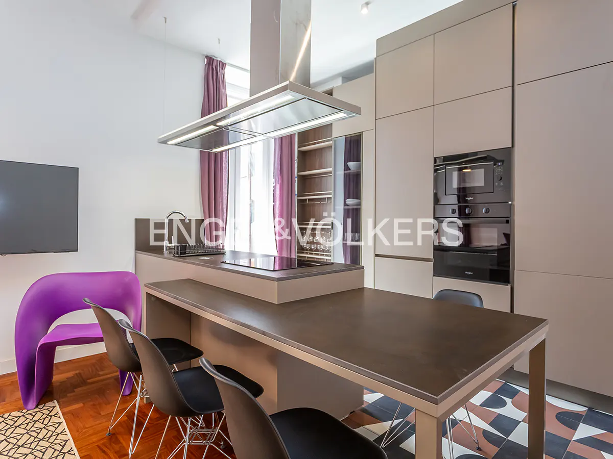 Modern kitchen with a dark countertop island, black chairs, and a purple chair. Stainless steel hood over the cooktop, and beige cabinets.