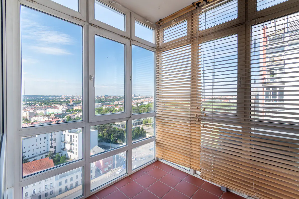 Enclosed balcony with red tile floor, white framed windows, and wood blinds. Cityscape visible through the windows.