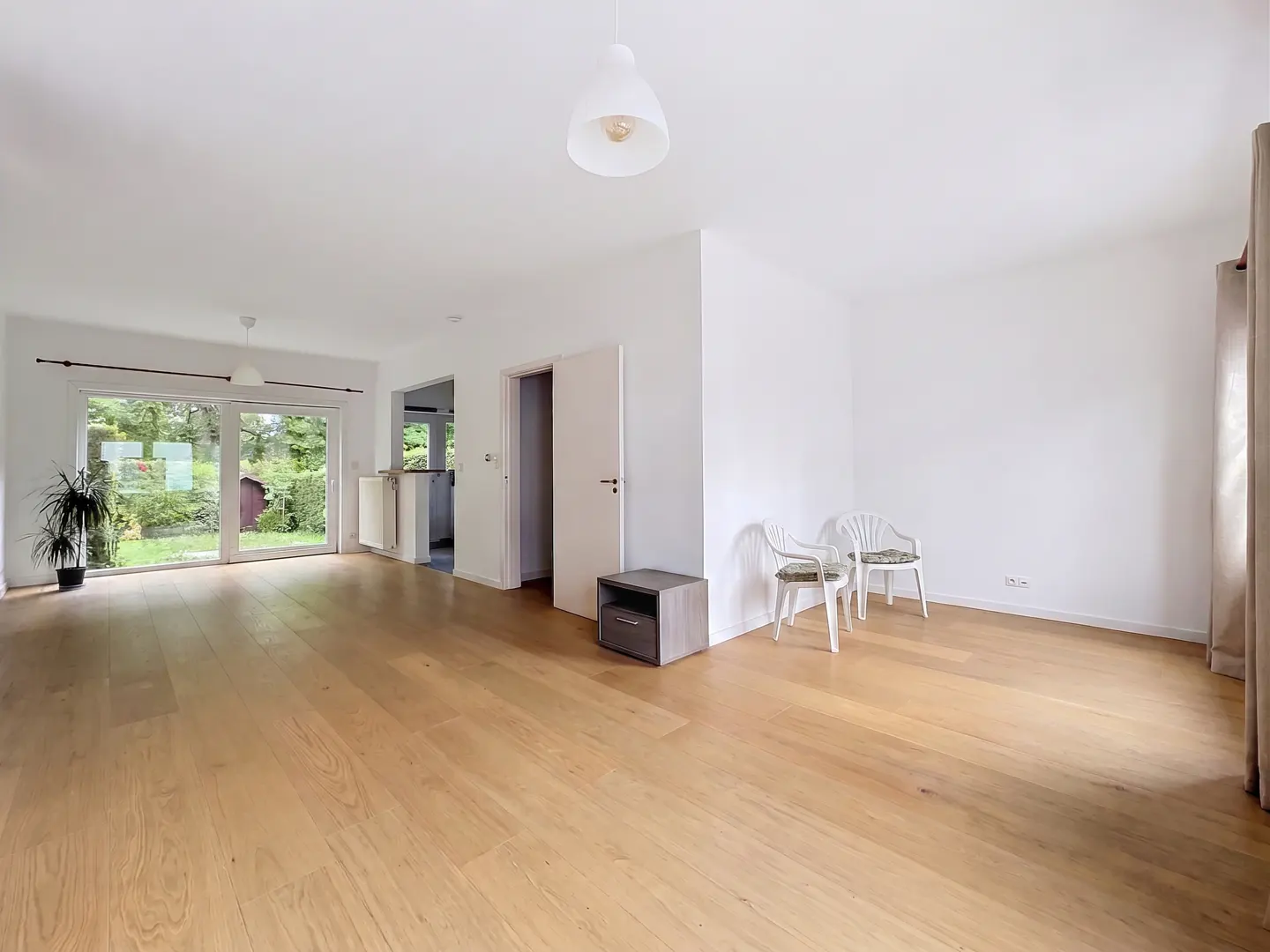 Bright, empty living room with wood floors, white walls, and a sliding glass door to a green yard. Two white chairs sit near a gray side table.