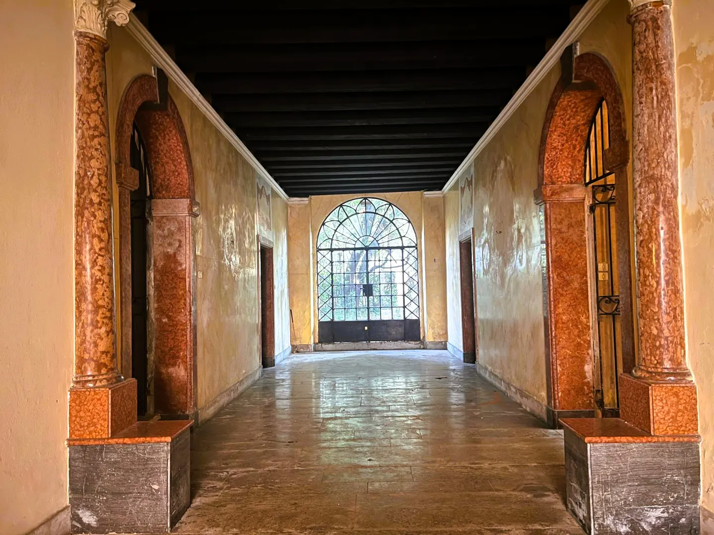 Hallway with marble columns and arches, leading to a large arched window with a black metal gate. Walls are yellow and worn.
