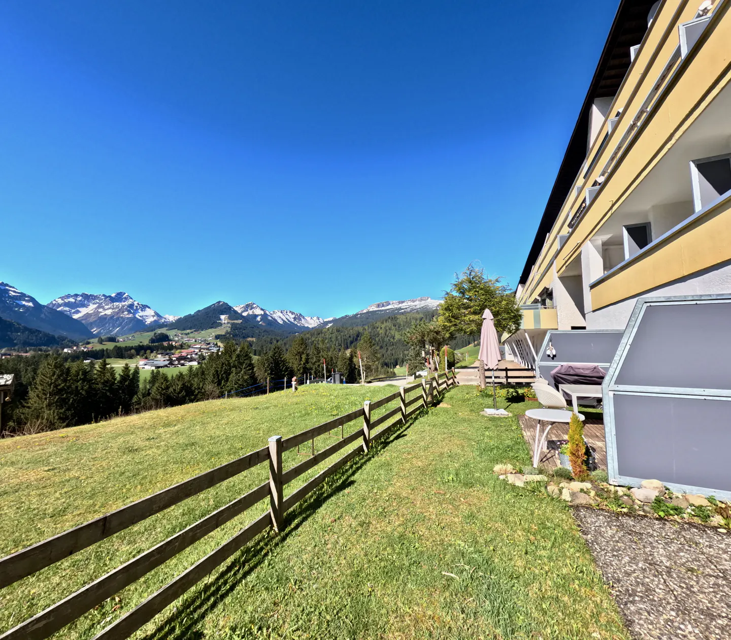 Scenic view of a yellow building with balconies overlooking a green lawn, wooden fence, and snow-capped mountains under a clear blue sky.