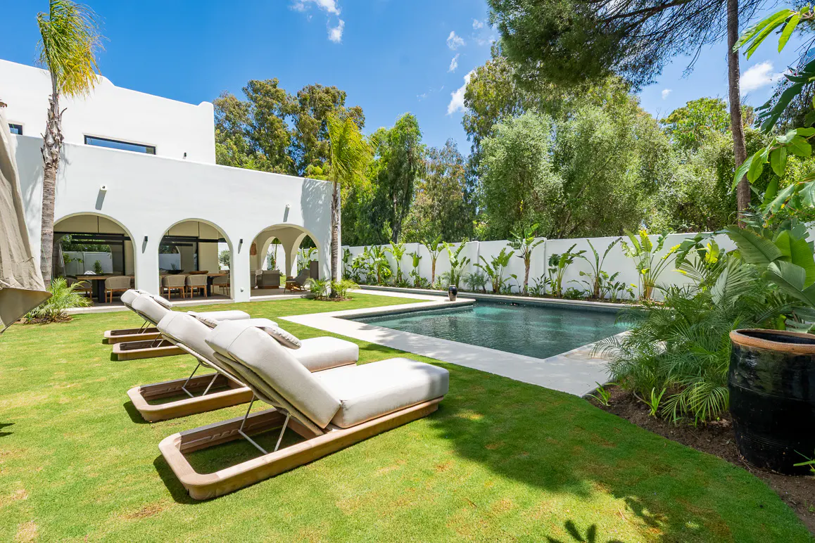Backyard view of a white house with a pool, lounge chairs, and green grass.