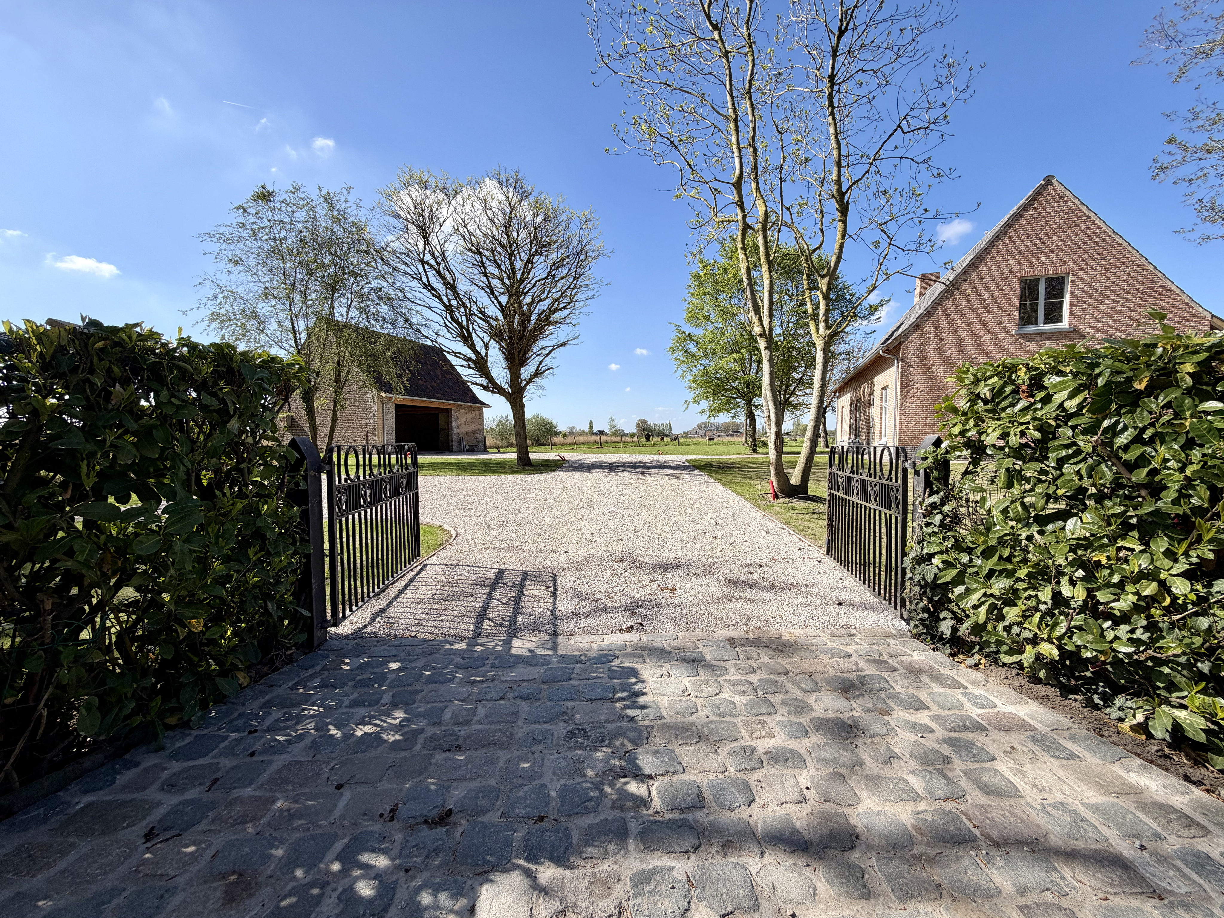 View of a gravel driveway leading to a brick house and barn, framed by an open black metal gate and green hedges.