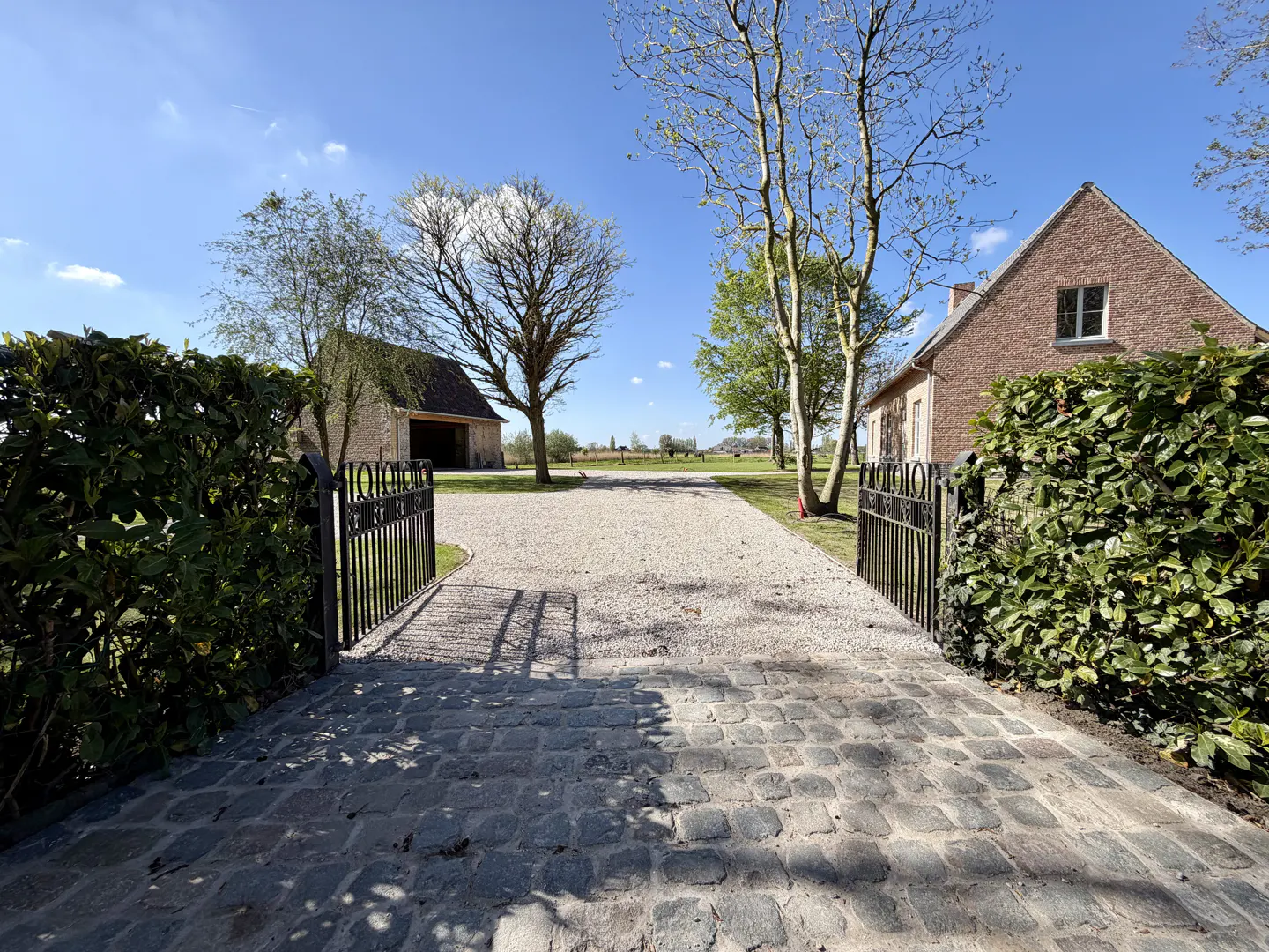 View of a gravel driveway leading to a brick house and barn, framed by an open black metal gate and green hedges.