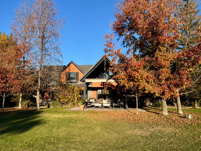 A two-story brick house with a black roof, surrounded by trees with autumn foliage, sits on a green lawn.