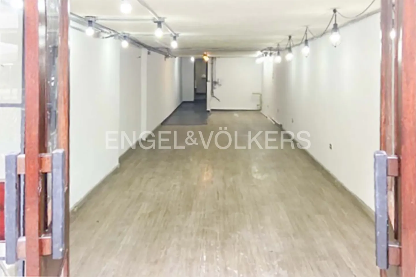 An open doorway reveals a long, empty commercial space with white walls, a light wood floor, and overhead lighting. The Engel & Völkers logo is centered in the image.
