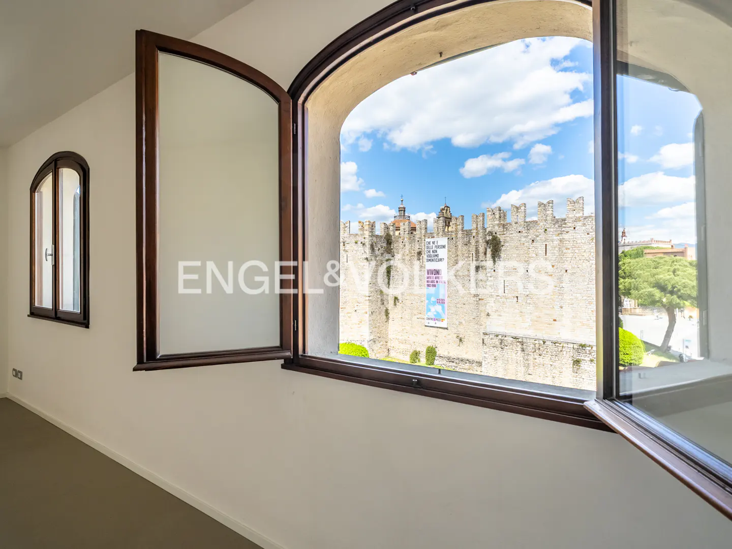 Interior view of a room with open arched windows, showcasing a castle under a blue sky.