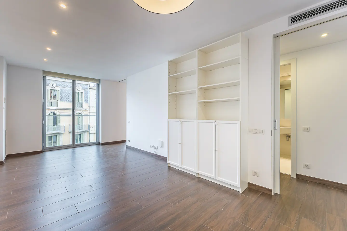 Bright, empty room with dark wood floors, white walls, and a large window. A white bookcase and open bathroom door are on the right.