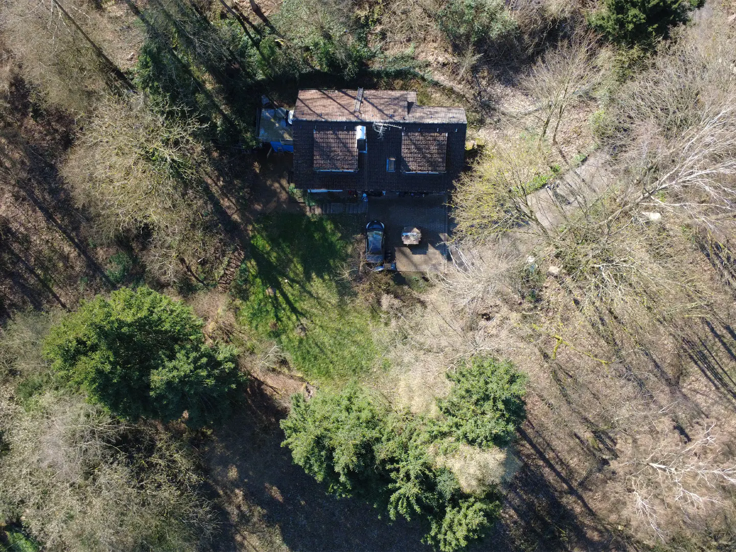 Aerial view of a house with a brown roof, surrounded by trees, a car and a stone table in the driveway.
