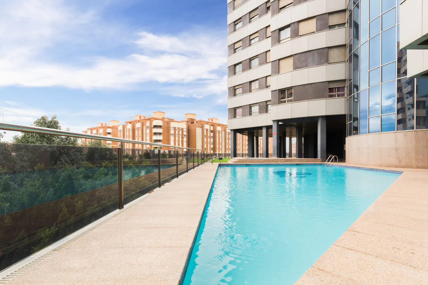 Outdoor pool with blue water next to a modern apartment building on a sunny day. Other buildings are visible in the background.