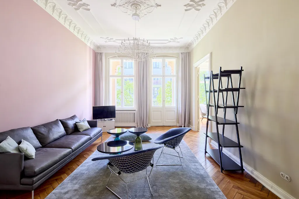 Living room with pink and beige walls, herringbone wood floors, grey rug, black sofa, and modern chairs. A chandelier hangs from the ornate ceiling.