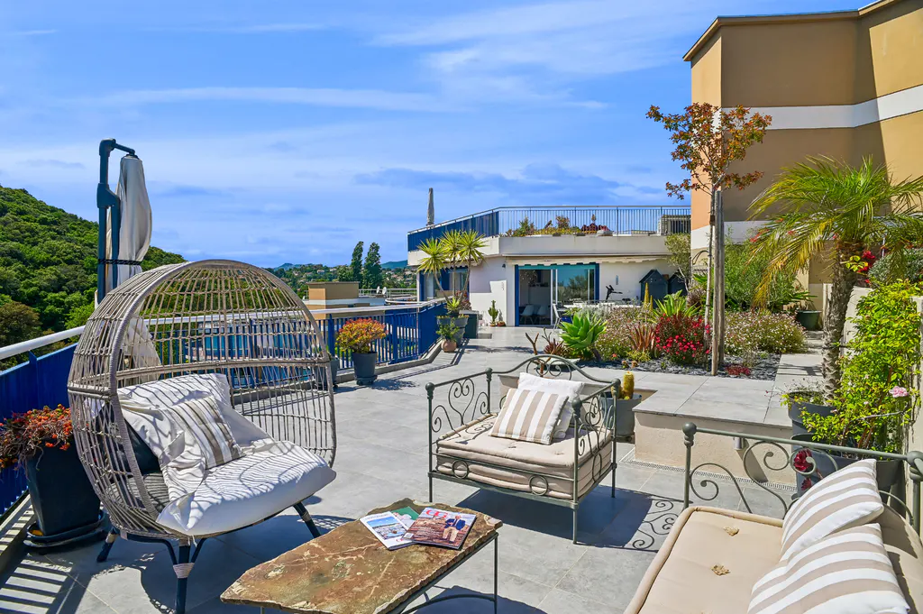 Rooftop patio with wicker chair, metal seating, and stone table with magazines. Lush greenery and blue sky in the background.